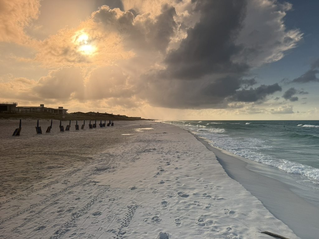 Sun burning gold through storm clouds over a wide, empty white sand beach with dune fencing