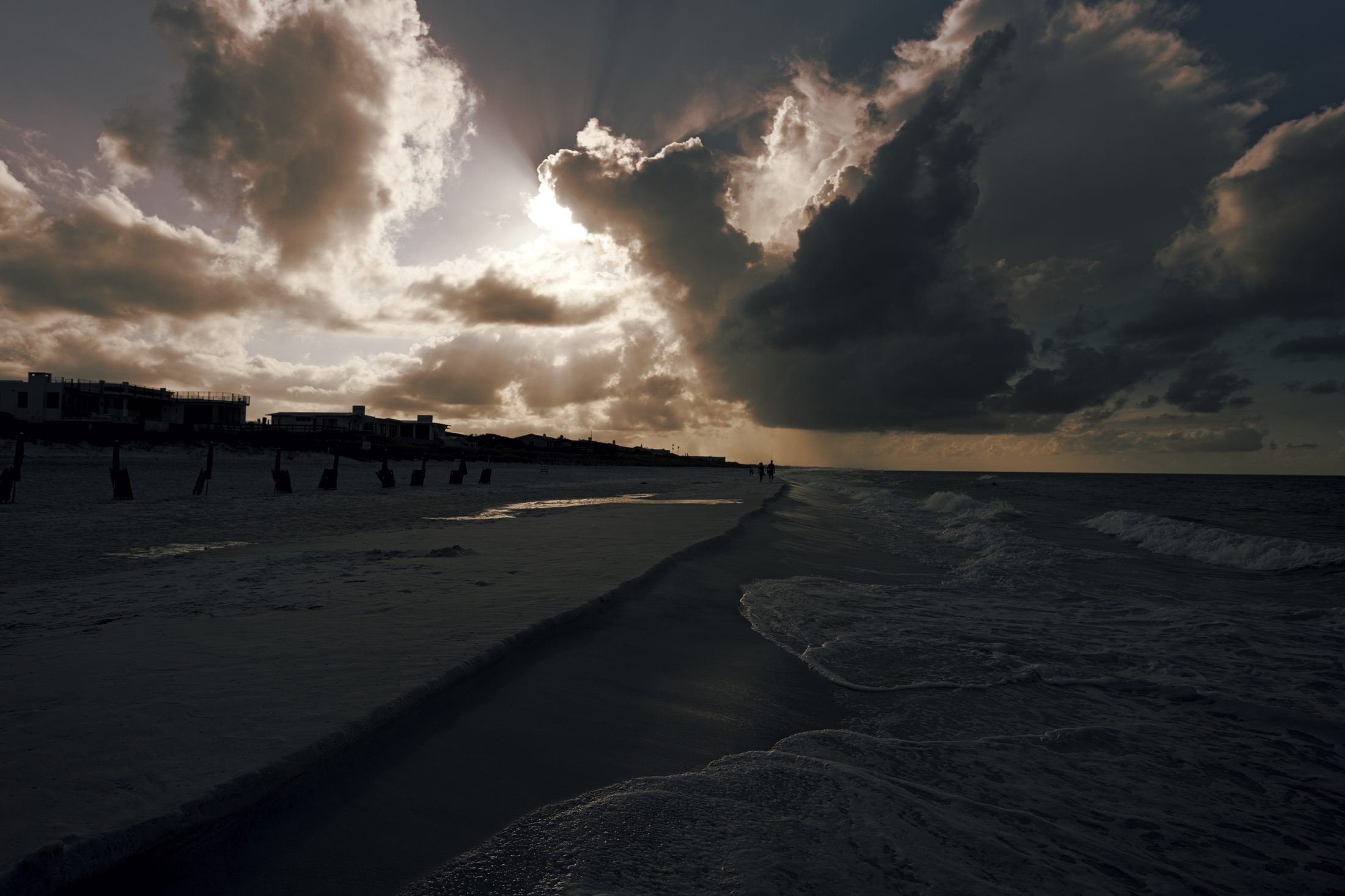 Dark storm front and sunburst over the empty beach, dune fence posts and rooflines in silhouette, two small figures at the waterline