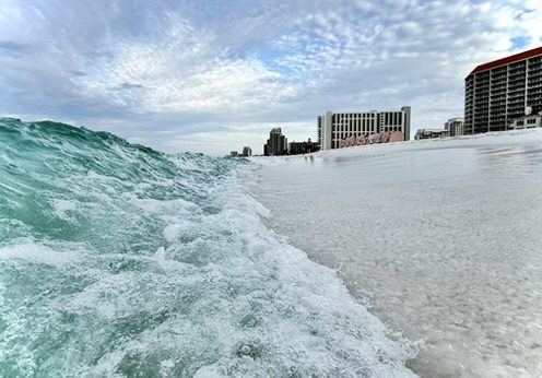 A green wave breaking in the foreground with high-rise towers along the beach behind it